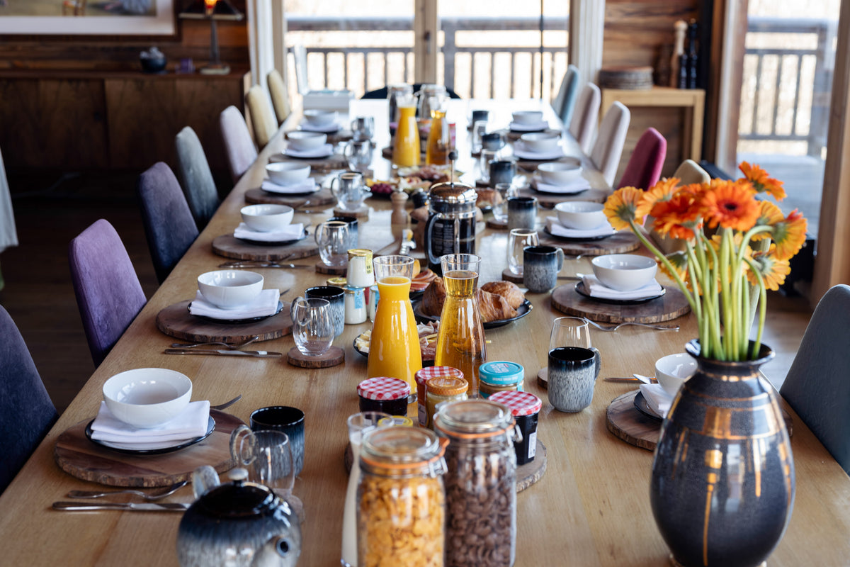 A long wooden dining table set for multiple guests in a rustic cabin-style dining room, decorated with white bowls, plates, glass mugs, jars, and a large vase of orange flowers.