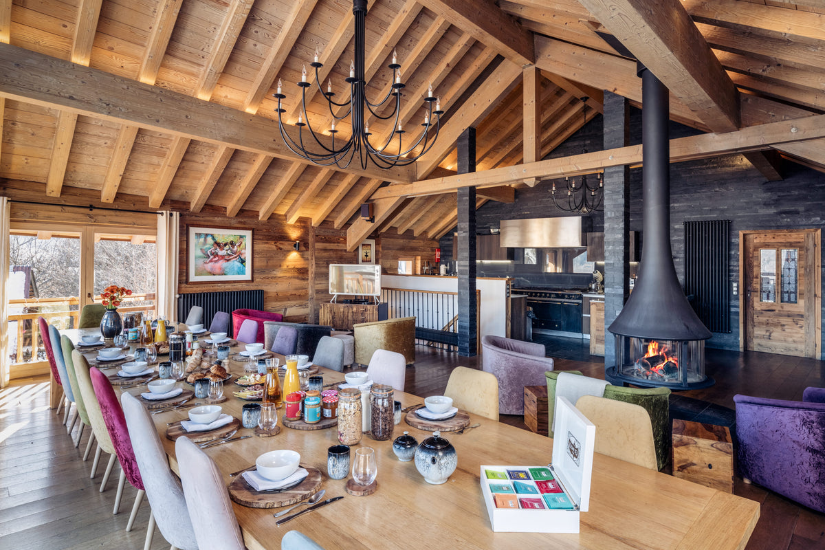 A rustic wooden dining room with a long table set for multiple guests, a large black pendant chandelier, a wood-burning fireplace with a chimney pipe, and colorful upholstered chairs in a mountain-lodge interior.