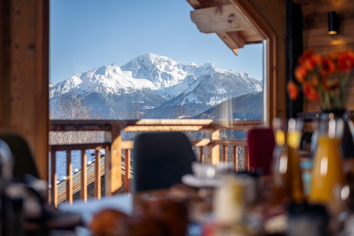 A scenic view from a rustic wooden chalet balcony looking out at snow-covered mountains under a clear blue sky, with blurred breakfast table details in the foreground.