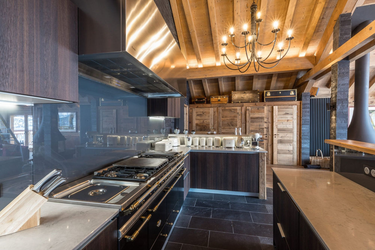 A luxury modern kitchen interior featuring dark wood cabinetry, a large stainless steel gas range, a black glass backsplash, a rustic wood ceiling with warm lighting, and a black chandelier.