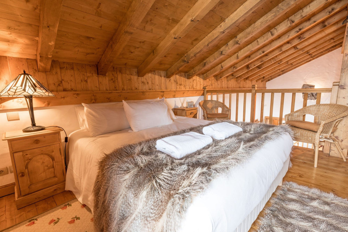 A cozy bedroom with a bed dressed in white linens and a gray faux-fur throw, set in a rustic wooden attic with a slanted ceiling, wooden beams, a bedside table with a lamp, and a wicker chair.