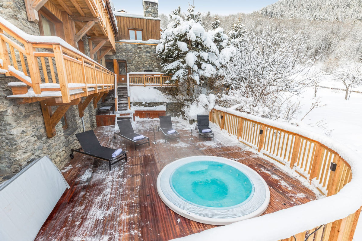 A luxury chalet outdoor winter scene featuring a round hot tub with turquoise water on a wooden deck, surrounded by snow-covered trees and mountains.