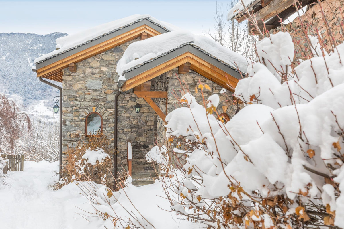 A stone chalet-style cabin with snow-covered roofs and wooden trim, surrounded by deep snow and leafless bushes in a winter mountain setting.