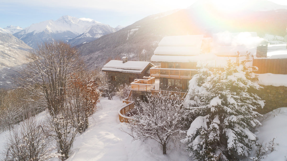 A snowy mountain alpine scene with a snow-covered chalet, bare trees, evergreen trees laden with snow, and distant peaks under bright sun with lens flare.