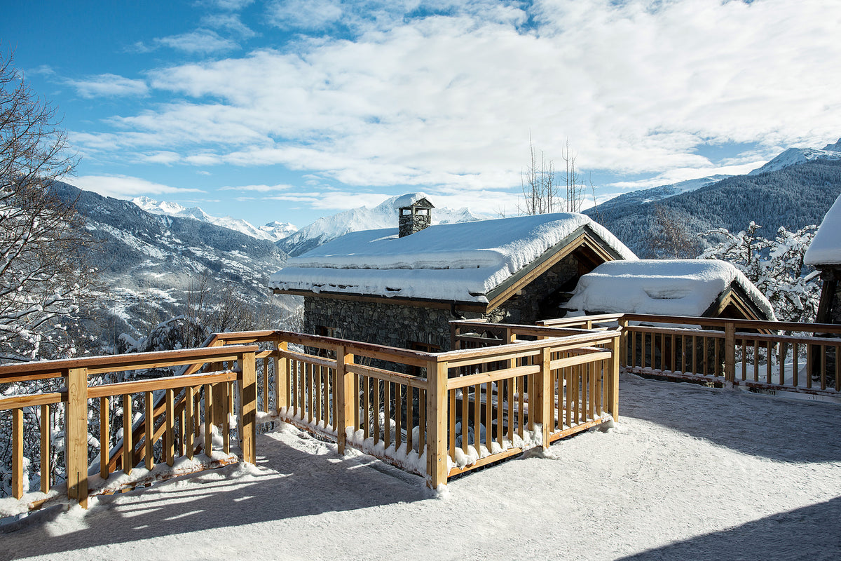 A snowy mountain chalet with wooden balconies overlooking a wide alpine valley, with snow-covered peaks and a blue sky with white clouds in the background.