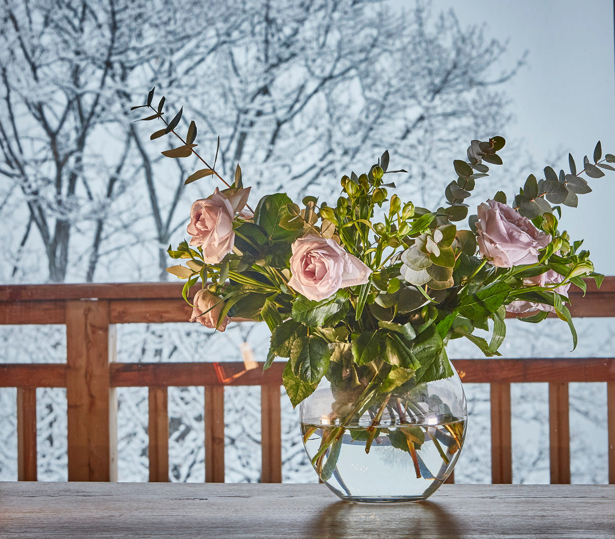A bouquet of pale pink roses and green foliage arranged in a clear glass vase on a wooden table, with a snowy outdoor railing and bare trees in the background.
