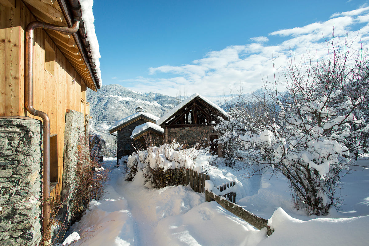 A snowy mountain village scene with wooden chalets, snow-covered trees, and distant mountains under a bright blue sky.