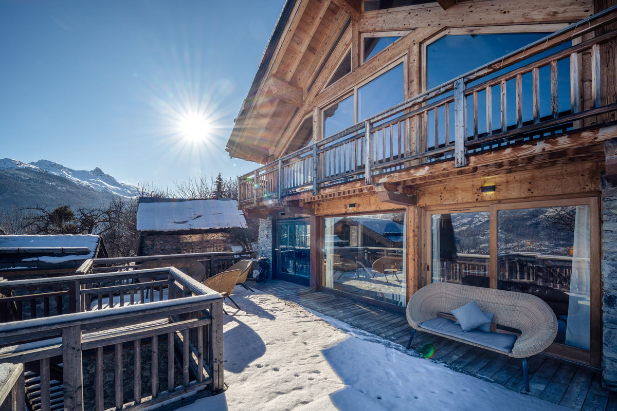 A sunny mountain chalet with wooden balconies, large glass windows, snow on the ground, and a wicker bench with pillows on the patio. Mountains and a blue sky with the sun are visible in the background.
