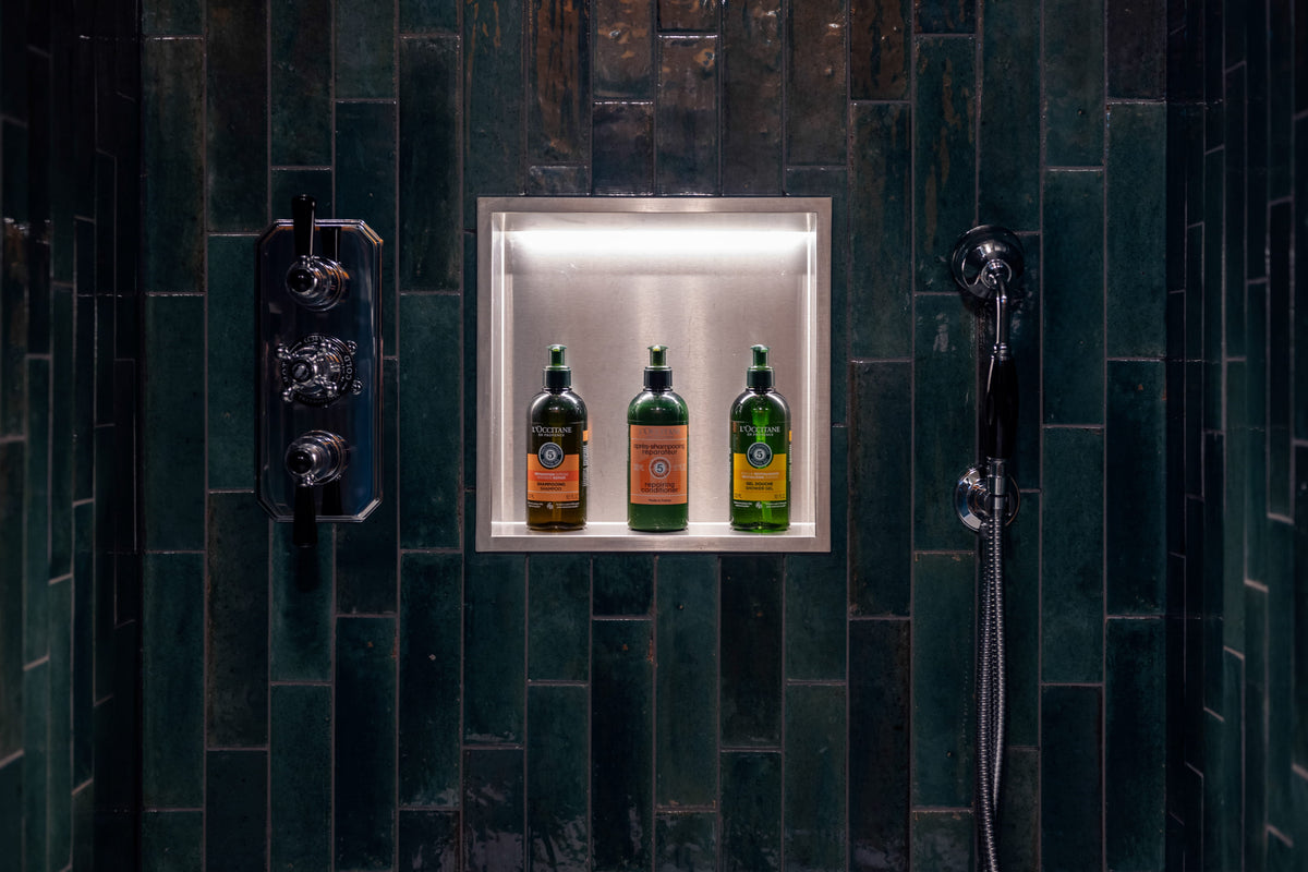 Three green bottles of Ginebreda liqueur with orange labels displayed in a built-in illuminated niche on a dark green tiled wall, flanked by chrome shower fixtures.