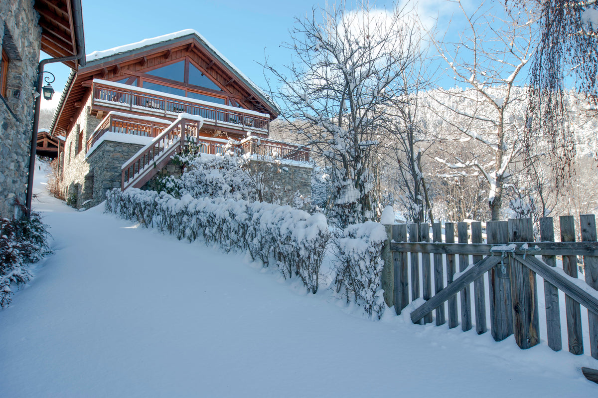 A snow-covered wooden chalet with a staircase leading up the slope, surrounded by snow-laden shrubs, bare winter trees, and a wooden fence under a blue sky.