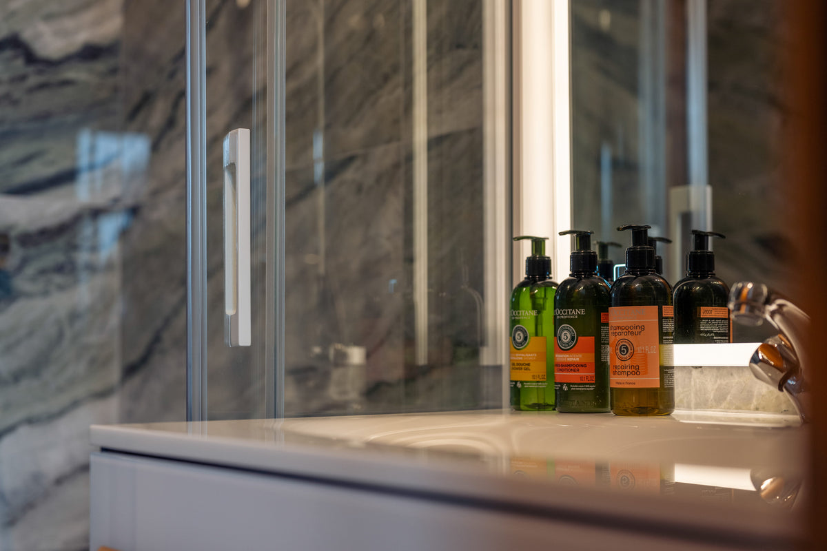 Four travel-sized toiletry bottles with black pumps and orange labels arranged on a bathroom counter in front of a glass shower door.
