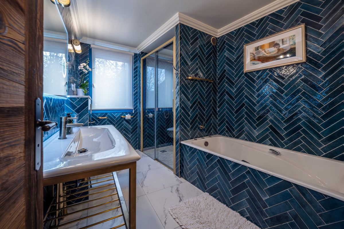 A modern bathroom interior with dark blue herringbone tile walls, a white bathtub, a white sink vanity, a glass shower enclosure, and a framed picture on the wall.