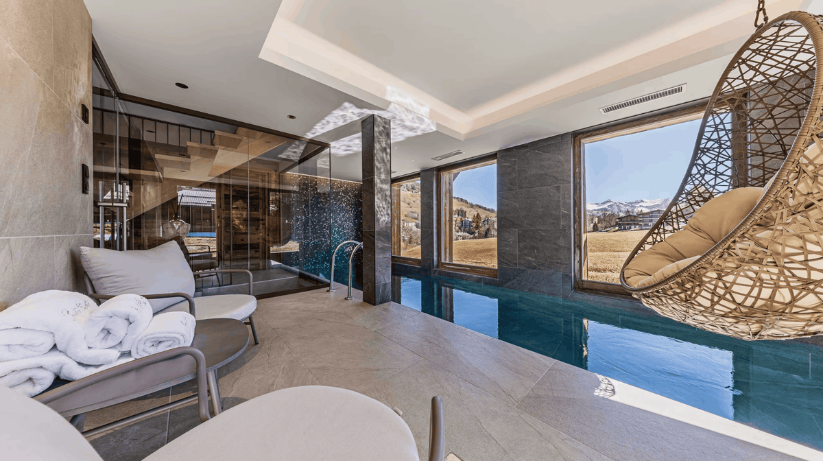 Luxury indoor poolside lounge area with a hanging woven egg chair, white towels, modern lounge seating, and large windows overlooking a mountain landscape.