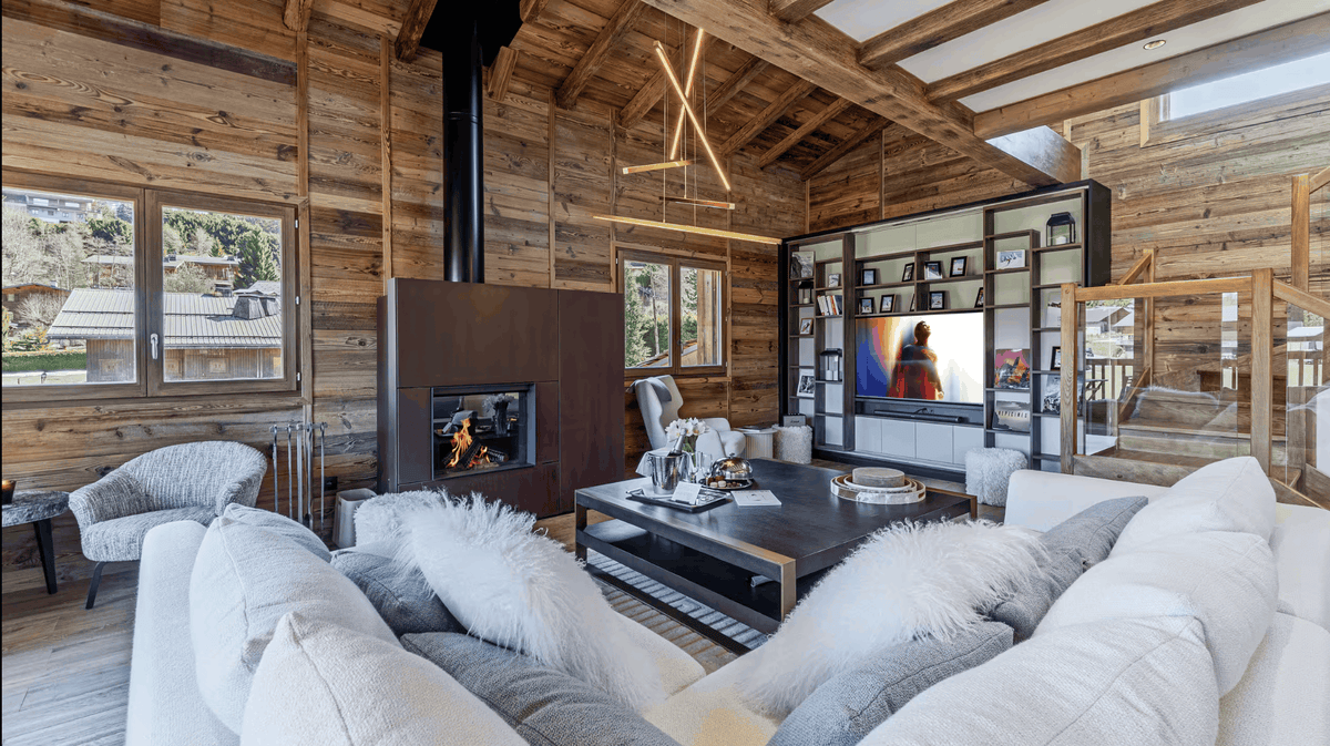 A modern luxury living room inside a rustic wooden chalet, featuring a large white sectional sofa with fluffy white and gray pillows, a dark wood coffee table, a black fireplace with a visible fire, and a large wall-mounted TV screen.