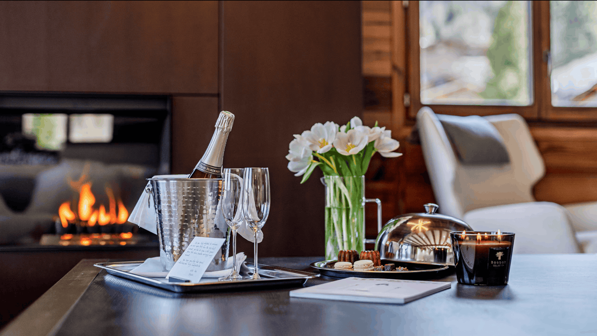A cozy indoor tabletop scene with a lit fireplace in the background. In the foreground, a silver ice bucket holds a champagne bottle, two champagne flutes, a glass vase with white flowers, a silver cloche, and a black candle jar with a visible 'VEDL' logo.