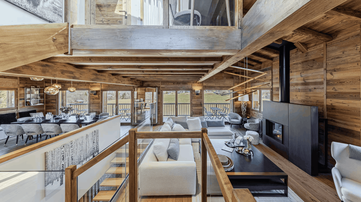 A modern luxury chalet interior featuring a high-ceilinged loft with exposed wood beams, a light gray sofa, a black coffee table, a large black fireplace, and a dining area in the background.