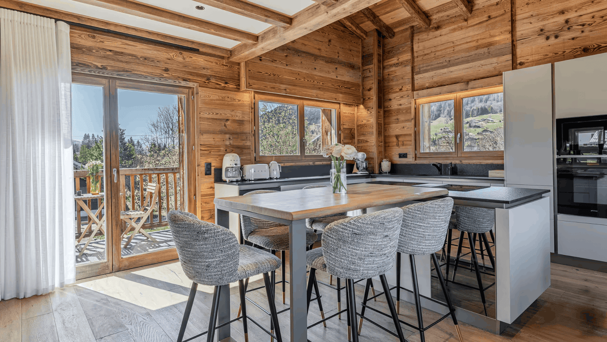 A modern rustic kitchen island with a wooden top and gray upholstered bar stools, set in a log-cabin style kitchen with wood-paneled walls and large windows.