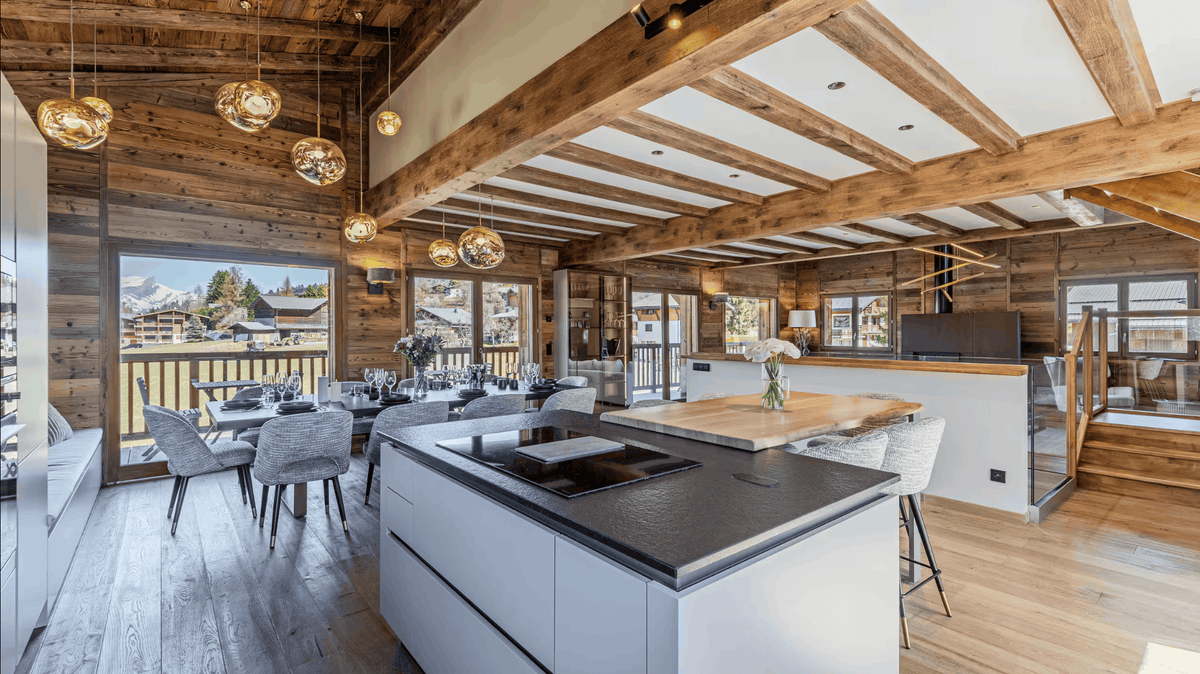 A luxury modern chalet interior featuring a large white kitchen island with a dark countertop, wooden ceiling beams, hanging glass globe pendant lights, and a dining area with mountain views.