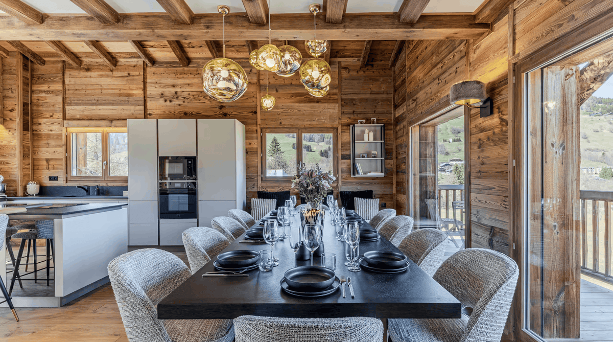A luxury modern dining room with a long dark wood table set with black plates and glassware, surrounded by woven gray chairs. The room features rustic wooden walls and ceiling, a kitchen island in the background, and large windows looking out onto a mountain landscape.