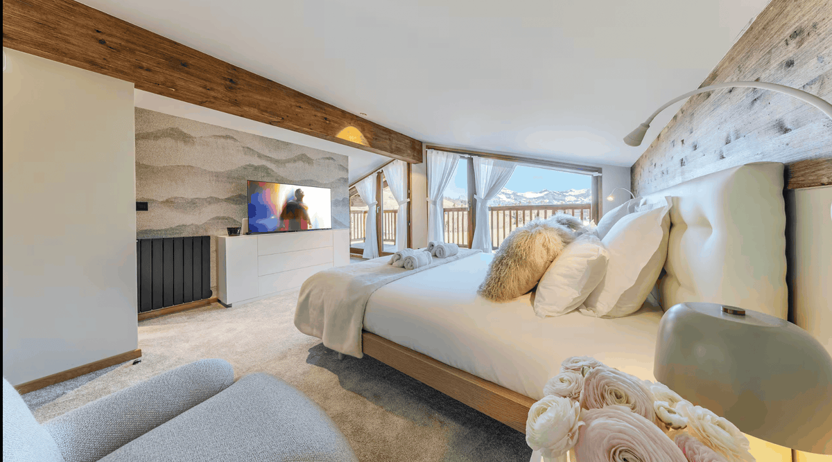 A modern ski chalet bedroom featuring a large bed with white bedding, plush pillows, and a textured beige headboard. The room has rustic wooden ceiling beams, a stone accent wall with a mounted TV, and a balcony with a mountain view in the background.
