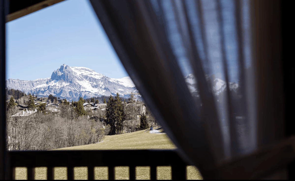 A scenic view of a snow-capped mountain peak above a grassy valley with trees and houses, framed by a dark wooden balcony railing and a sheer curtain in the foreground.