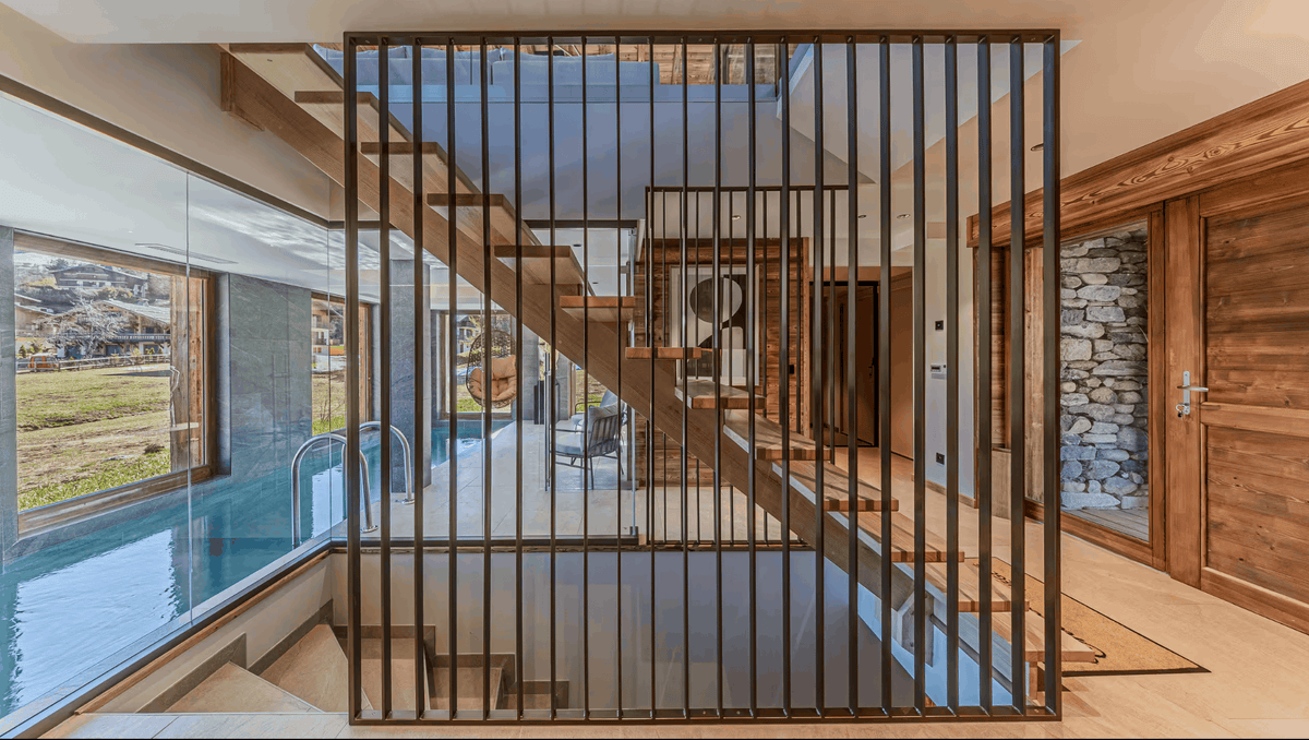 A modern interior architectural scene with a large black metal slat screen divider in the foreground, wooden staircase, glass walls, stone wall, and pool area visible through large windows.