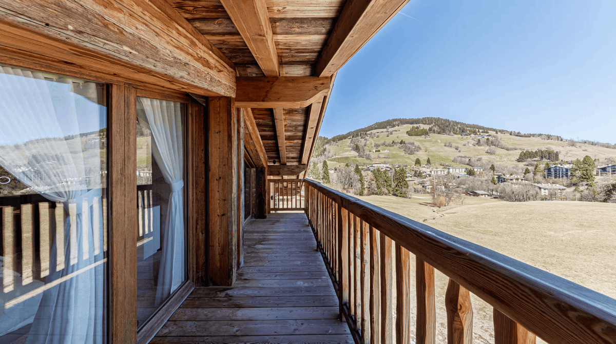 A wooden balcony with a railing overlooking a mountain valley landscape under a clear blue sky.