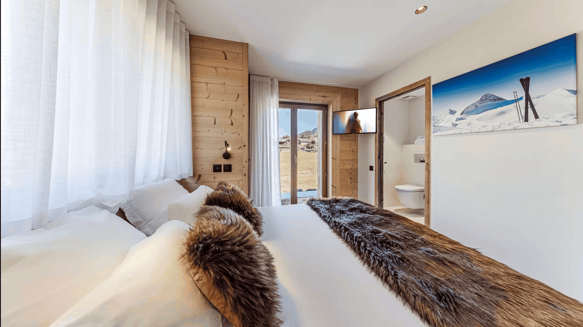 A modern bedroom interior with a large bed dressed in white bedding and a dark brown faux-fur runner, set in a bright room with wood paneling, sheer curtains, and a framed mountain landscape print on the wall.