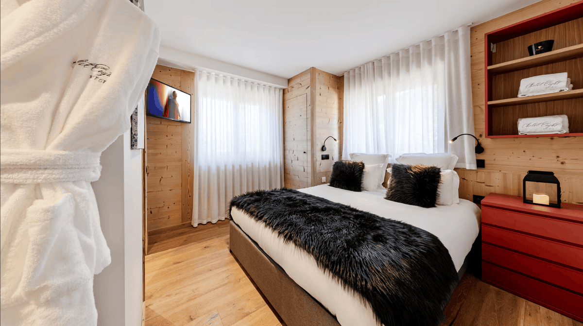 A modern bedroom interior with a neatly made bed, white bedding, black accent pillows, a black faux-fur throw, light wood walls and floor, white sheer curtains, a red side cabinet, built-in shelving, and a white robe hanging in the foreground.