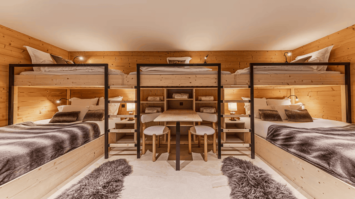 A modern ski chalet-style bedroom featuring three wooden bunk beds with black metal frames, arranged around a central wooden desk and two matching chairs. The room has warm wood-paneled walls, cozy bedside lamps, and plush gray faux-fur rugs on the floor.