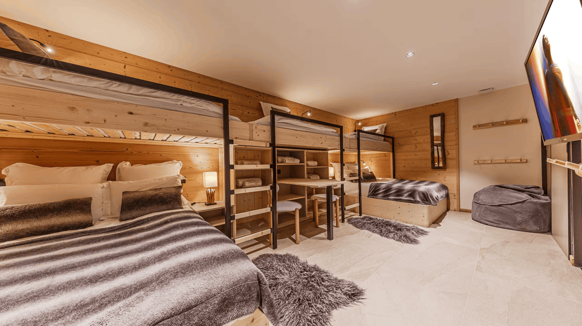 A modern ski resort style bedroom featuring two loft beds with black metal frames, wooden headboards, built-in shelving, a desk, and a flat-screen TV mounted on a wood-paneled wall.
