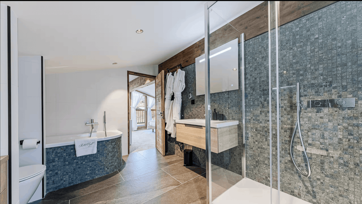 A modern luxury bathroom interior featuring a walk-in glass shower enclosure with a dark mosaic tile wall, a floating wooden vanity with a white sink, a bathtub with blue mosaic tile surround, and a walk-in closet with white robes.
