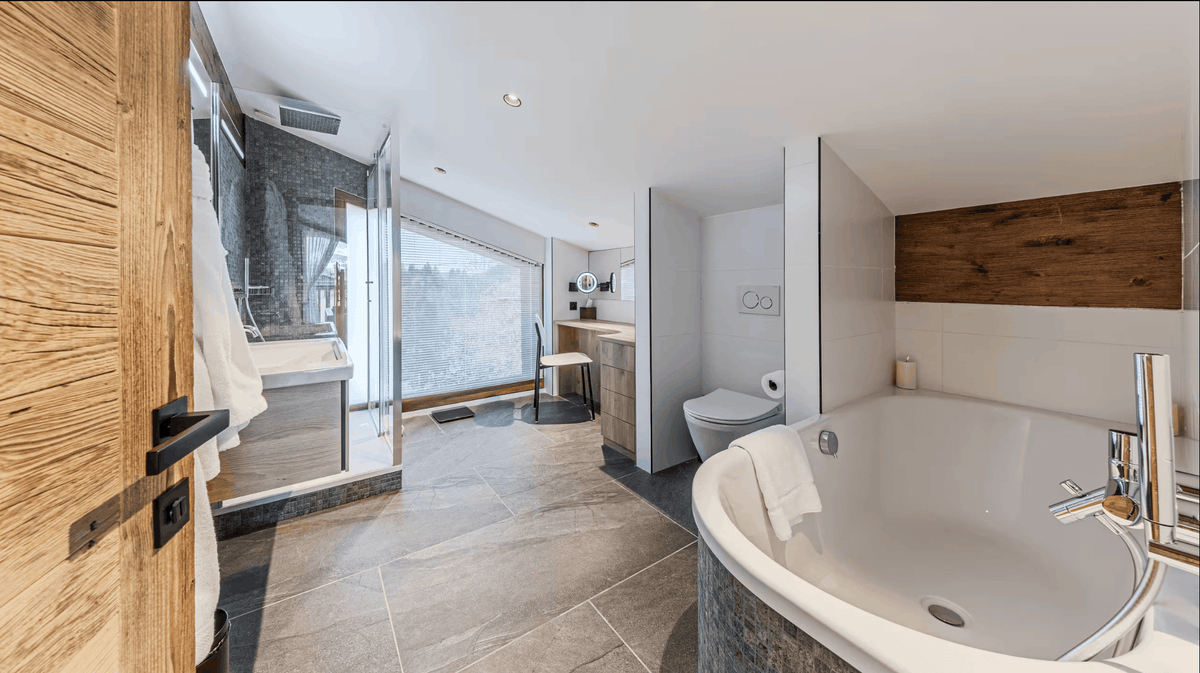 A modern luxury bathroom featuring a large white oval bathtub in the foreground, gray stone tile flooring, a glass shower enclosure, and a wooden door on the left.