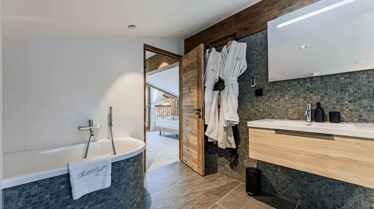 A luxury modern bathroom featuring a white bathtub with a chrome faucet, a light wood vanity with a white sink, a dark gray mosaic tile backsplash, and two white bathrobes hanging on a wooden door frame.