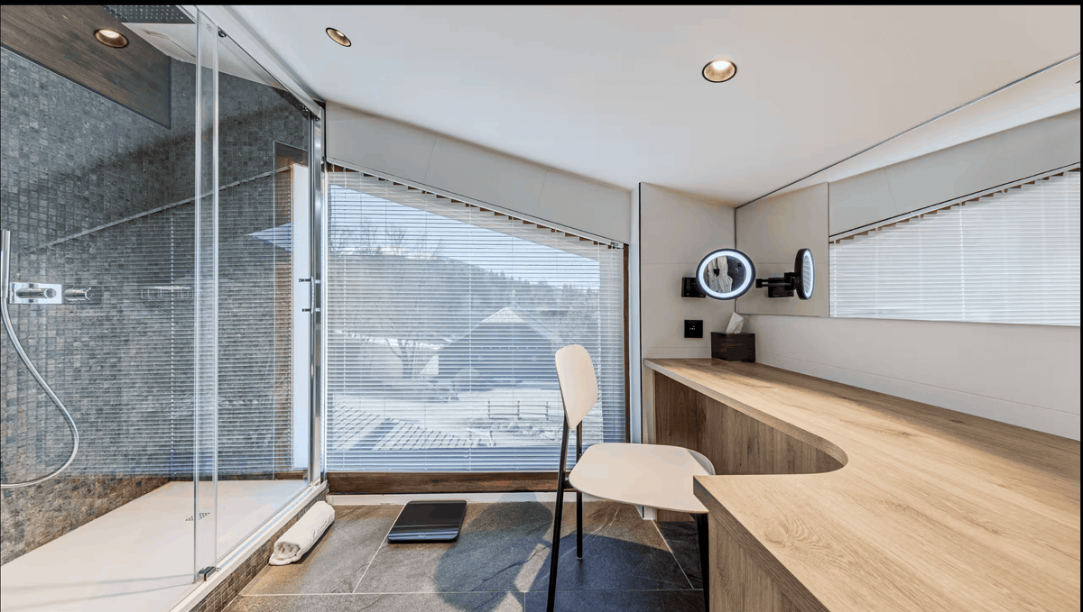 A modern bathroom interior with a glass shower enclosure, large window blinds, a light wood vanity desk, a white chair, and a round wall mirror.