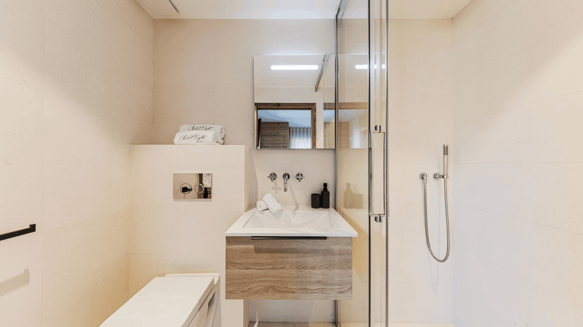 A modern bathroom vanity with a white sink, a light wood drawer cabinet, and a large mirror, set in a bright beige-toned bathroom with a glass shower enclosure.