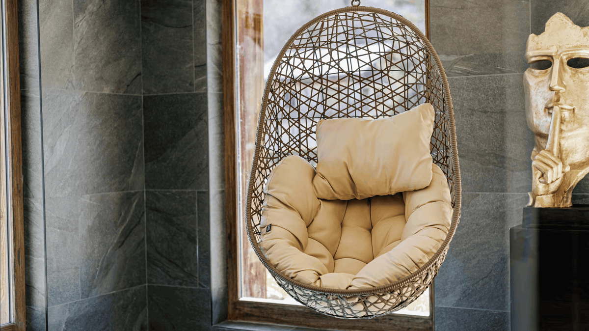 A hanging egg-shaped wicker chair with beige cushions, positioned in front of a window in a modern interior with dark gray stone tiles and a decorative gold bust on the right.