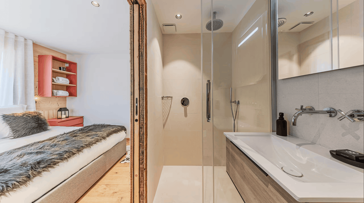 A modern bathroom interior with a walk-in shower, white sink vanity, mirrored wall, and an open doorway leading to a bedroom with a bed, wood shelving, and gray fur throw.