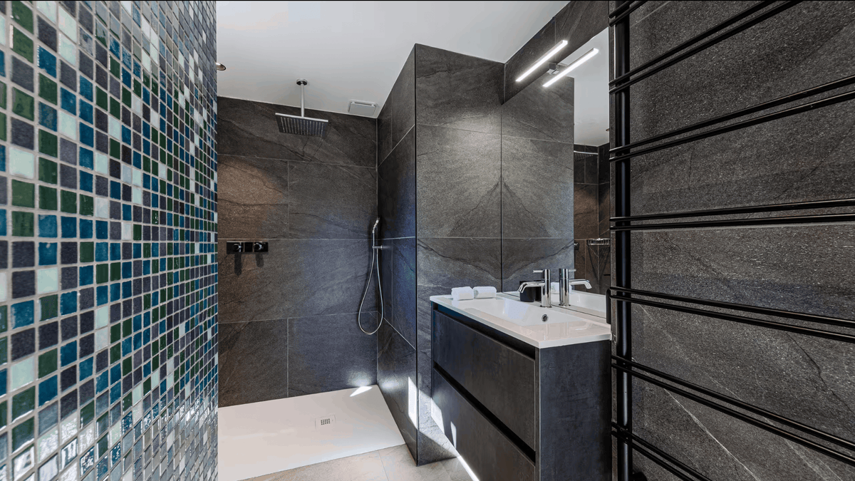 A modern bathroom interior with a walk-in shower, white sink vanity, mirrored wall, and an open doorway leading to a bedroom with a bed, wood shelving, and gray fur throw.