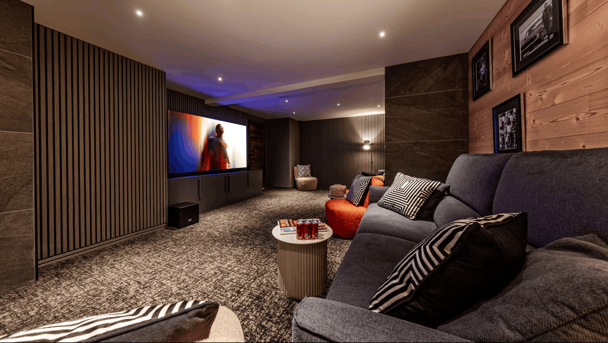 A modern home theater or living room setup with a large projection screen displaying a movie, upholstered gray sofas with striped and solid pillows, a small round coffee table, wood-paneled walls, and recessed ceiling lights.
