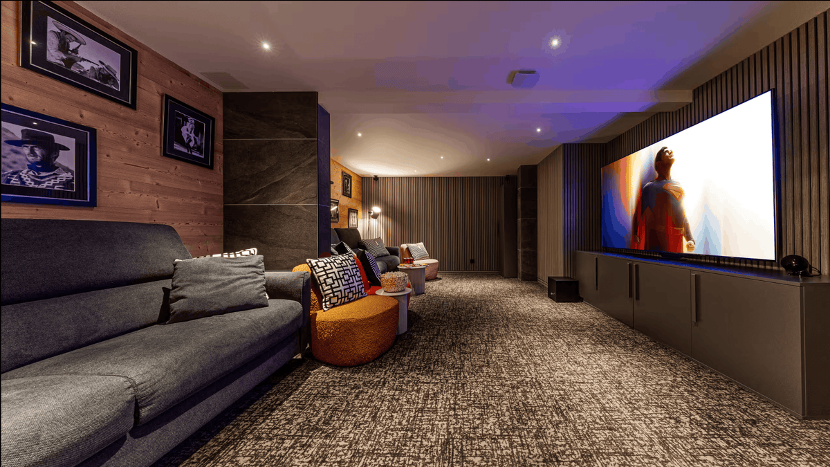 A modern home theater or living room setup with a large gray sofa, textured carpet, wall art, and a large illuminated screen displaying a movie scene. The room features wood-paneled walls, recessed ceiling lights, and ambient purple lighting.