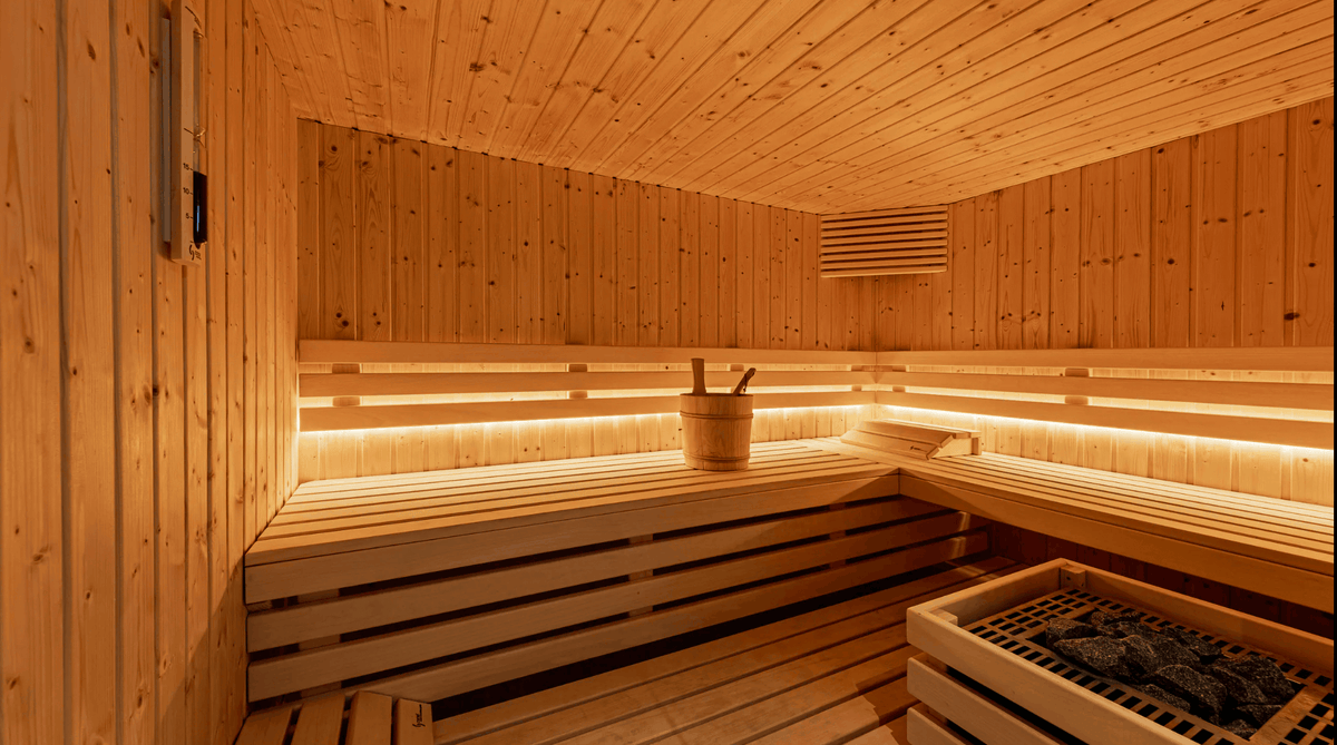 A warm wooden sauna interior with tiered seating, built-in LED lighting, a bucket with a wooden spoon on a bench, and a heating box with stones in the corner.