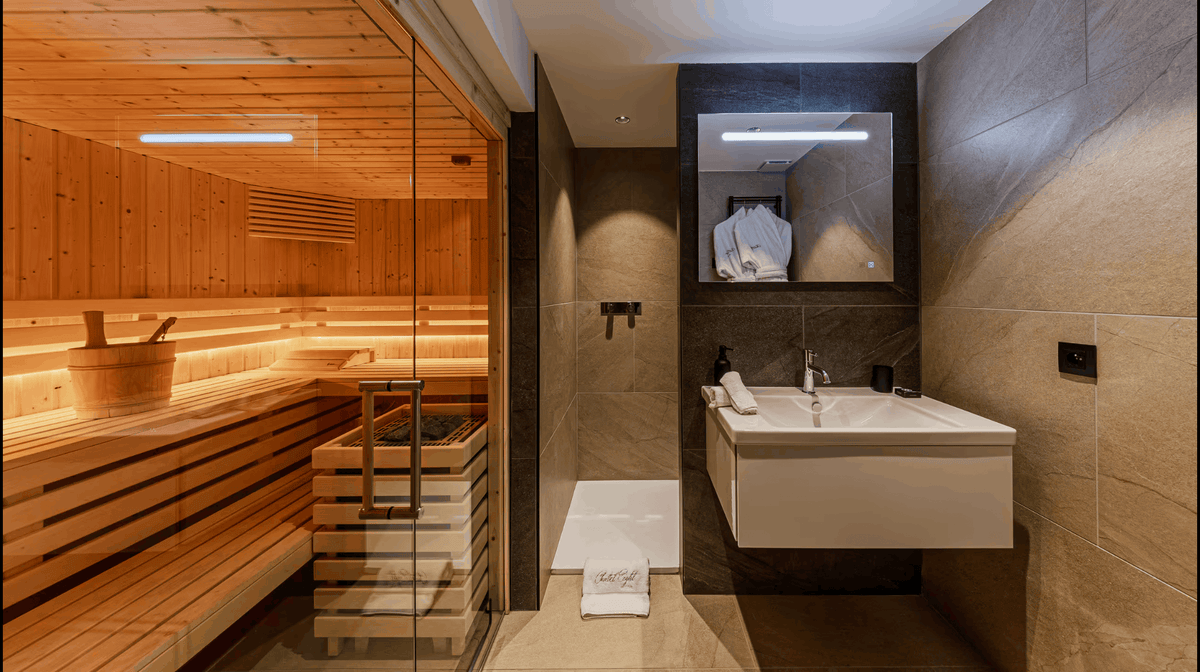 A modern bathroom interior with a glass-walled wooden sauna on the left, a white floating vanity sink on the right, and warm stone tile walls and floor.