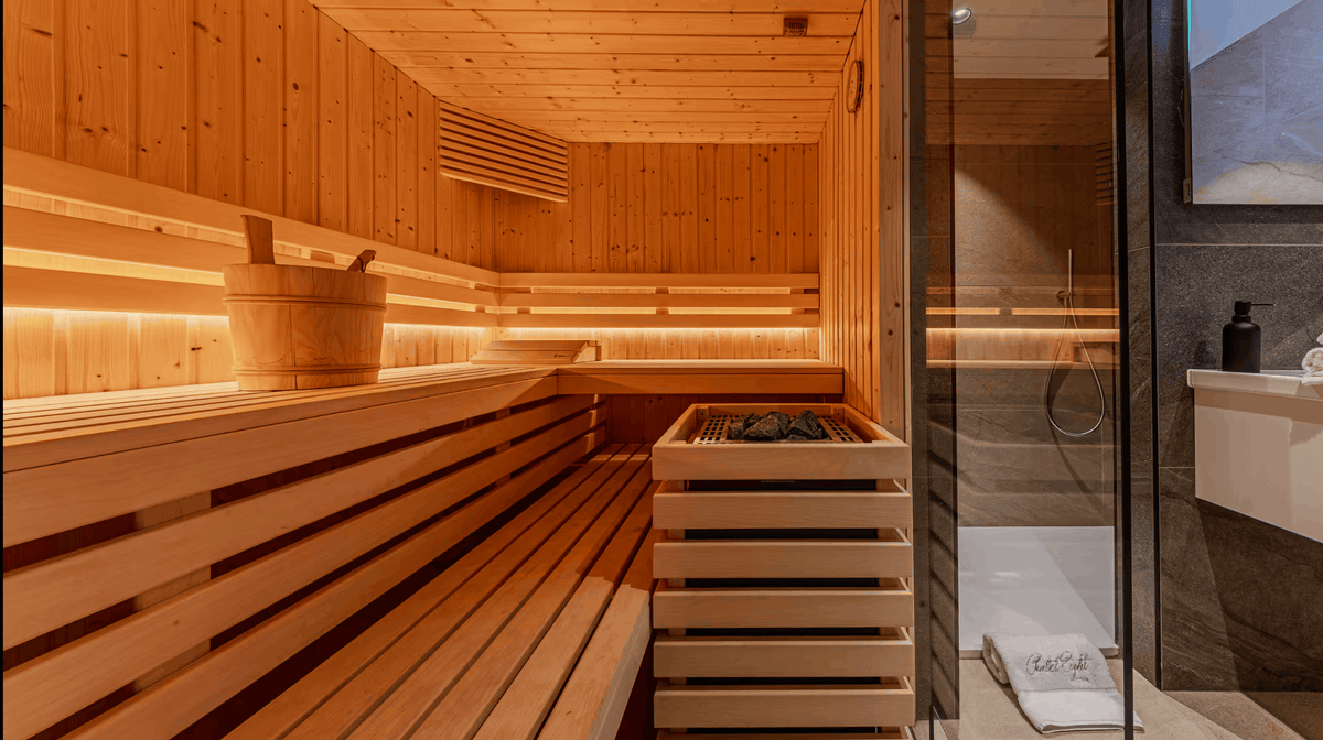 A warm wooden sauna interior with tiered benches, a bucket, a stacked sauna stone heater, and a glass door opening to a modern bathroom area.