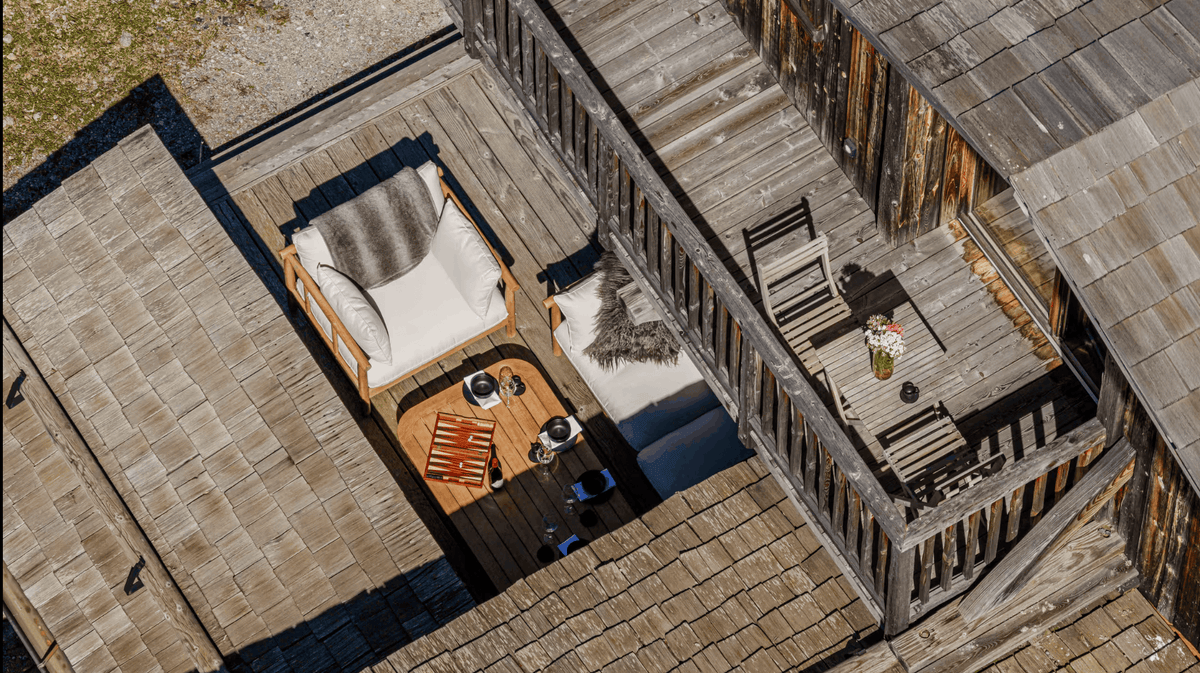 Aerial view of a rustic wooden cabin patio with a small seating area featuring a cushioned bench, a wooden coffee table, and a chair on a weathered deck surrounded by shingled roofs.