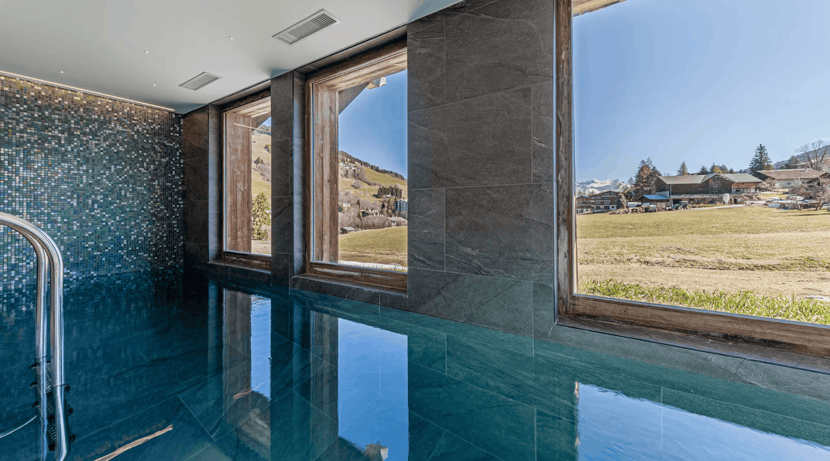 An indoor pool with deep blue water and dark stone walls, featuring large windows that offer a scenic view of a grassy field and mountains under a clear blue sky.