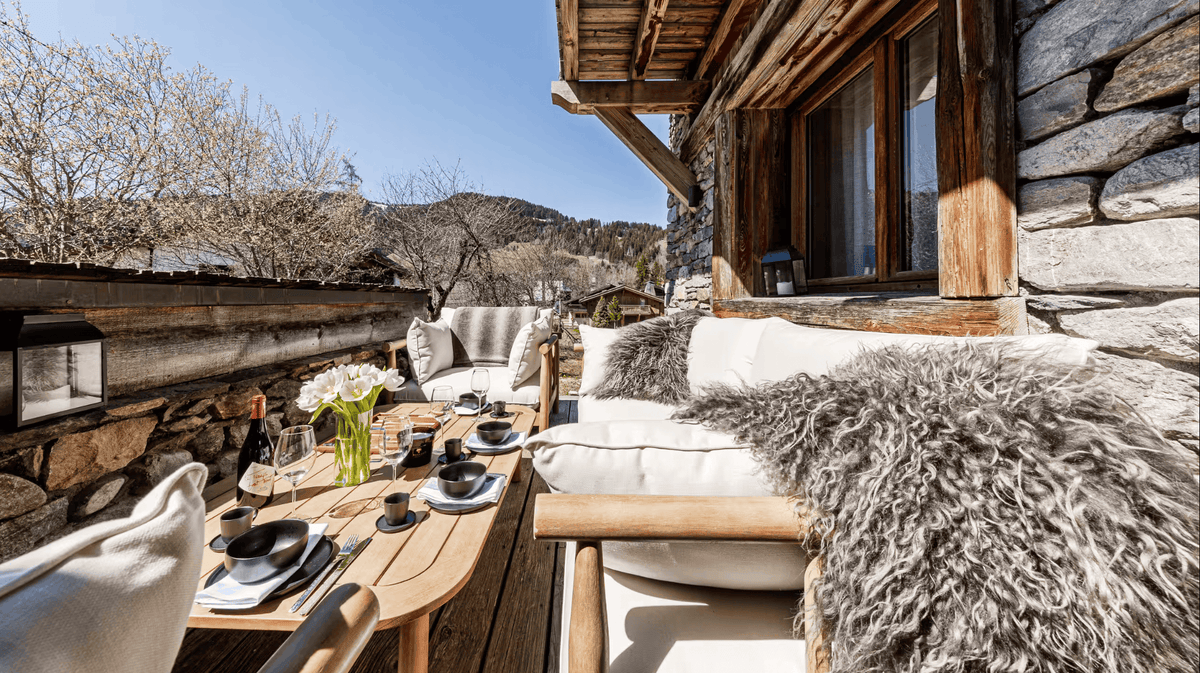 Outdoor patio seating area on a rustic wooden chalet deck, featuring light wood dining tables, cream cushions, gray faux-fur throw pillows, and a stone wall background with mountains and trees under a clear blue sky.