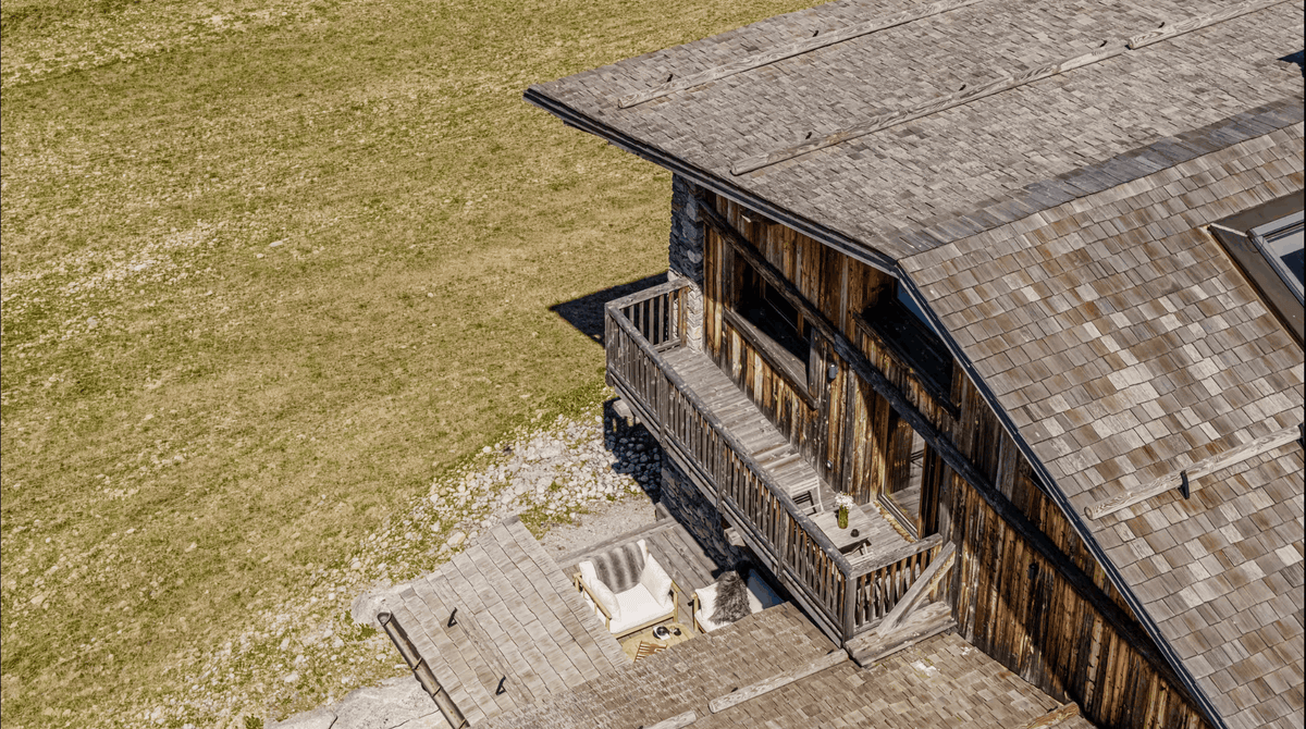 Aerial view of a rustic wooden chalet with a gray shingled roof, wooden siding, and a small patio area, set against a grassy field.