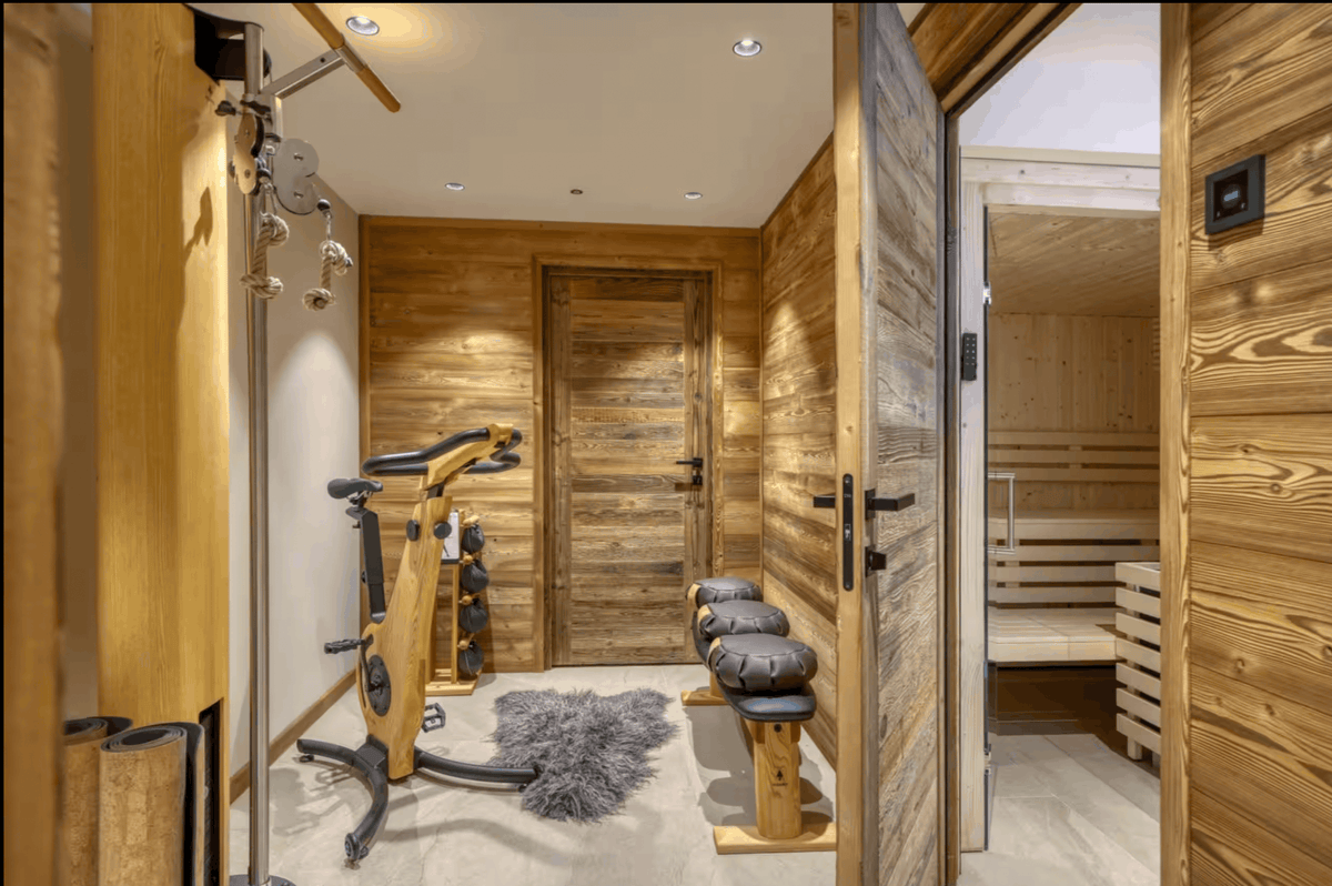A modern wooden sauna and gym room interior with a freestanding exercise bike, wooden benches, a shag rug, and an open door revealing a wooden sauna interior.