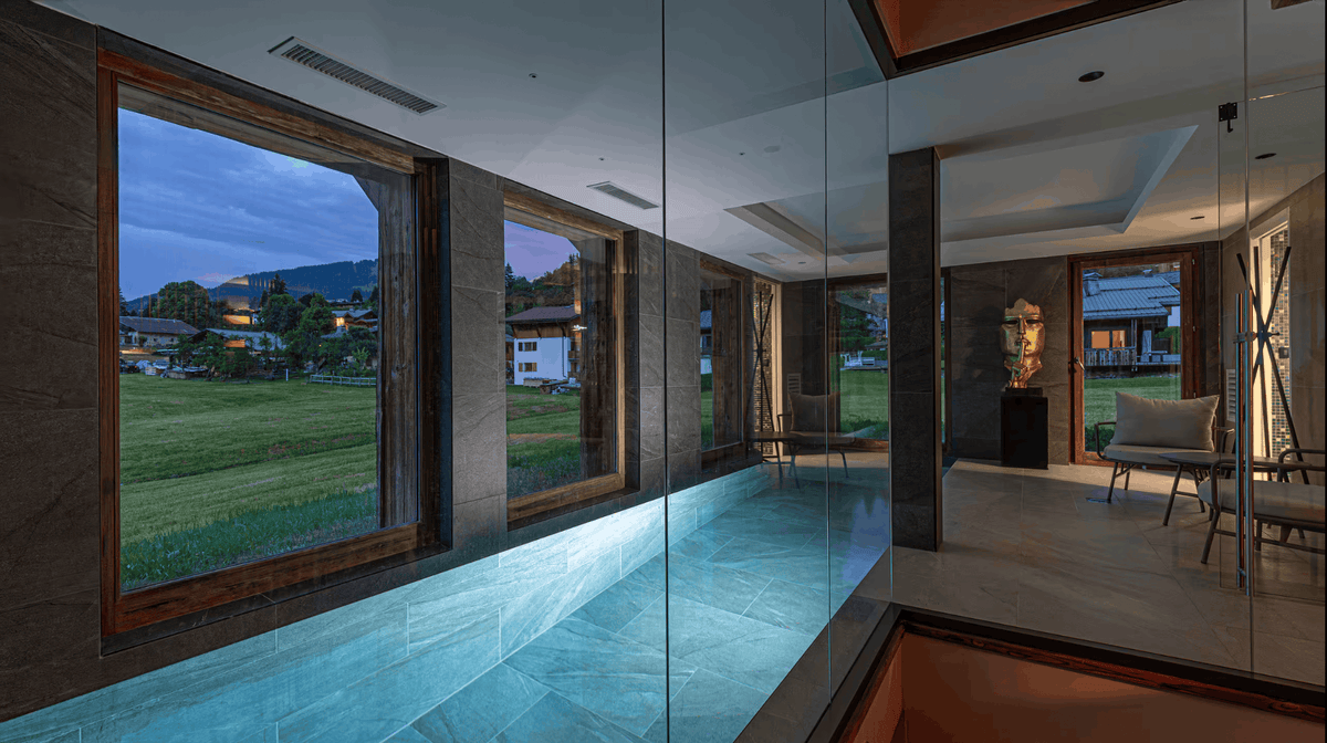 A modern indoor pool or walk-in bathtub with large floor-to-ceiling windows overlooking a grassy outdoor landscape at dusk. The space features dark wood framing, glass panels, and warm ambient lighting.